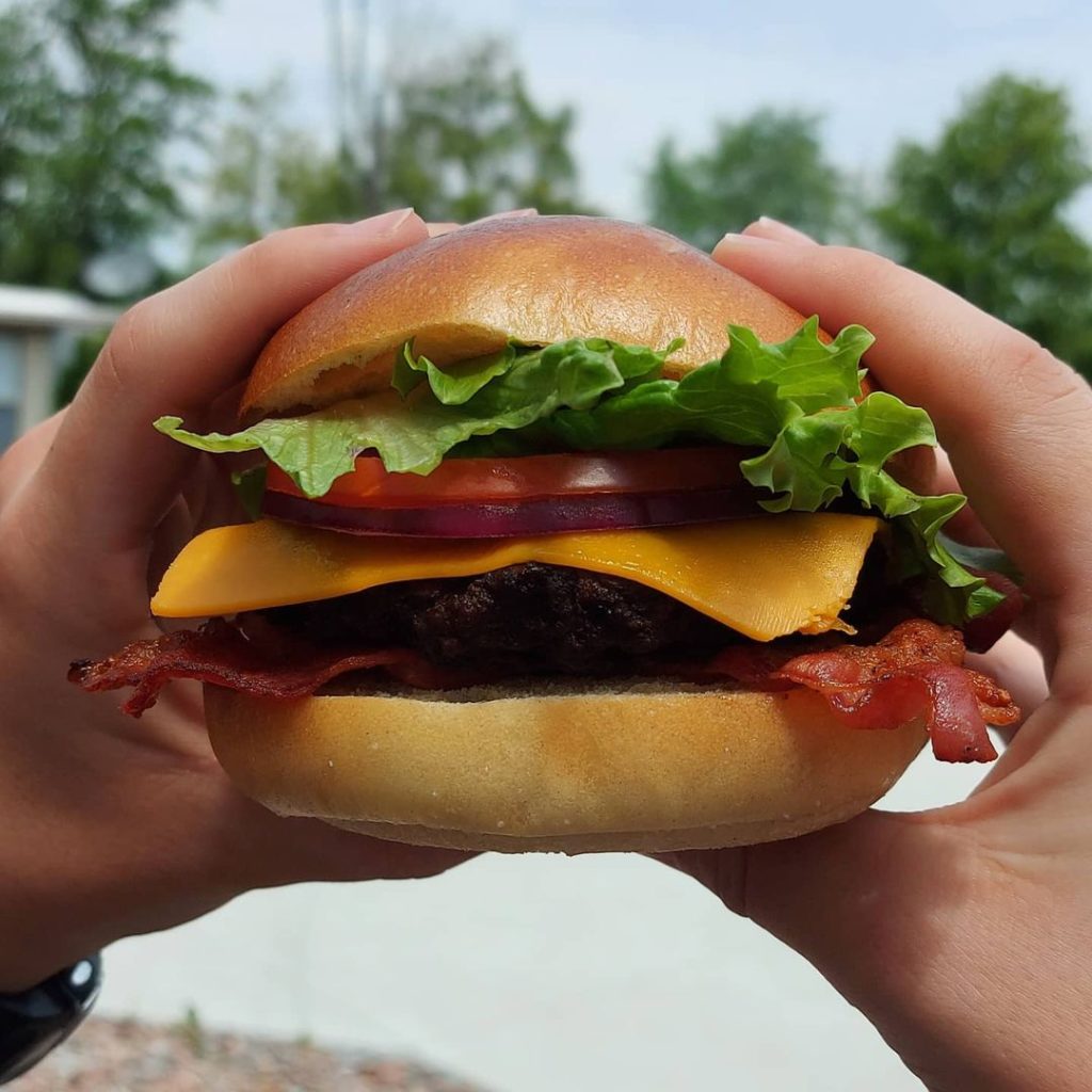 Two hands hold a delicious burger topped with bacon, cheese, purple onion, lettuce and tomato