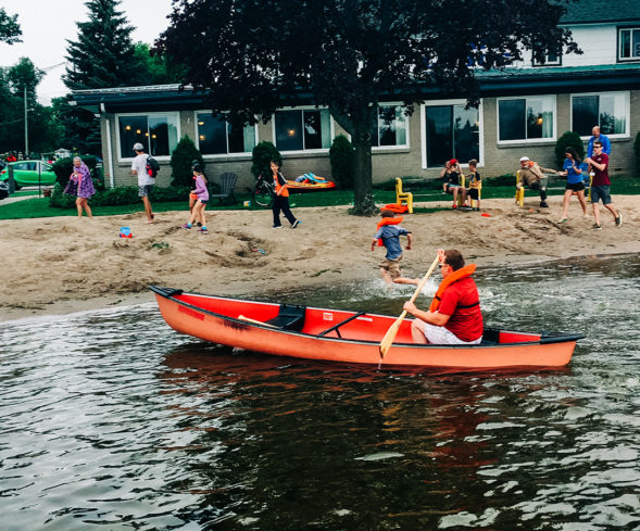 People enjoy the sandy beach at Elim Lodge while a man paddles by in a red canoe