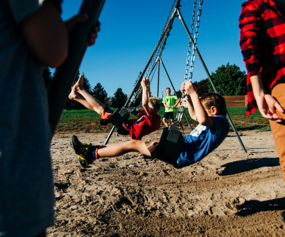 Children enjoy the swings at Elim Lodge playground while a volunteer staff member keeps watch